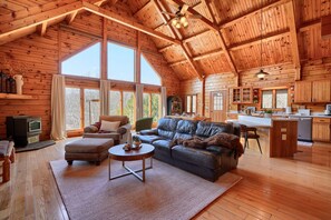 A spacious cabin interior with a high ceiling, wooden walls, a cozy seating area with a leather sofa and ottoman, a fireplace, and a kitchen area. - Waters Edge, Cozy Cabin & Pond (Creola)