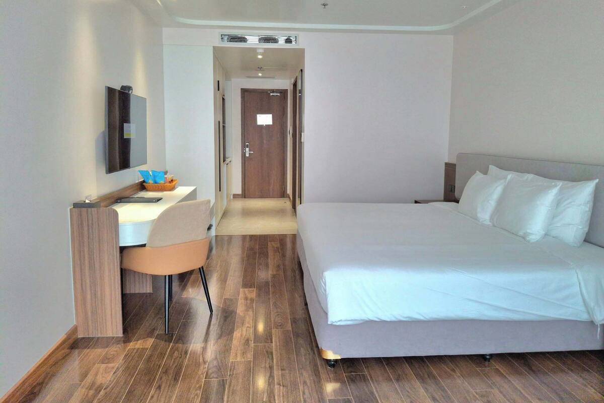 A hotel room with a bed, a desk with a chair, a TV mounted on the wall, and a wooden floor.