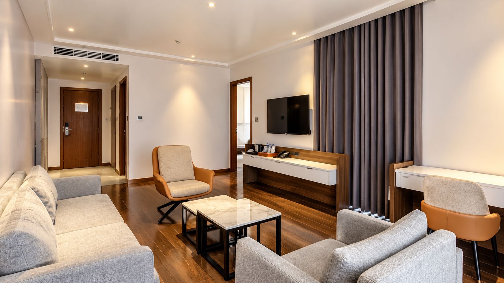 A modern hotel room with a sofa set, a coffee table, a desk with a flat-screen TV, and a built-in bench.