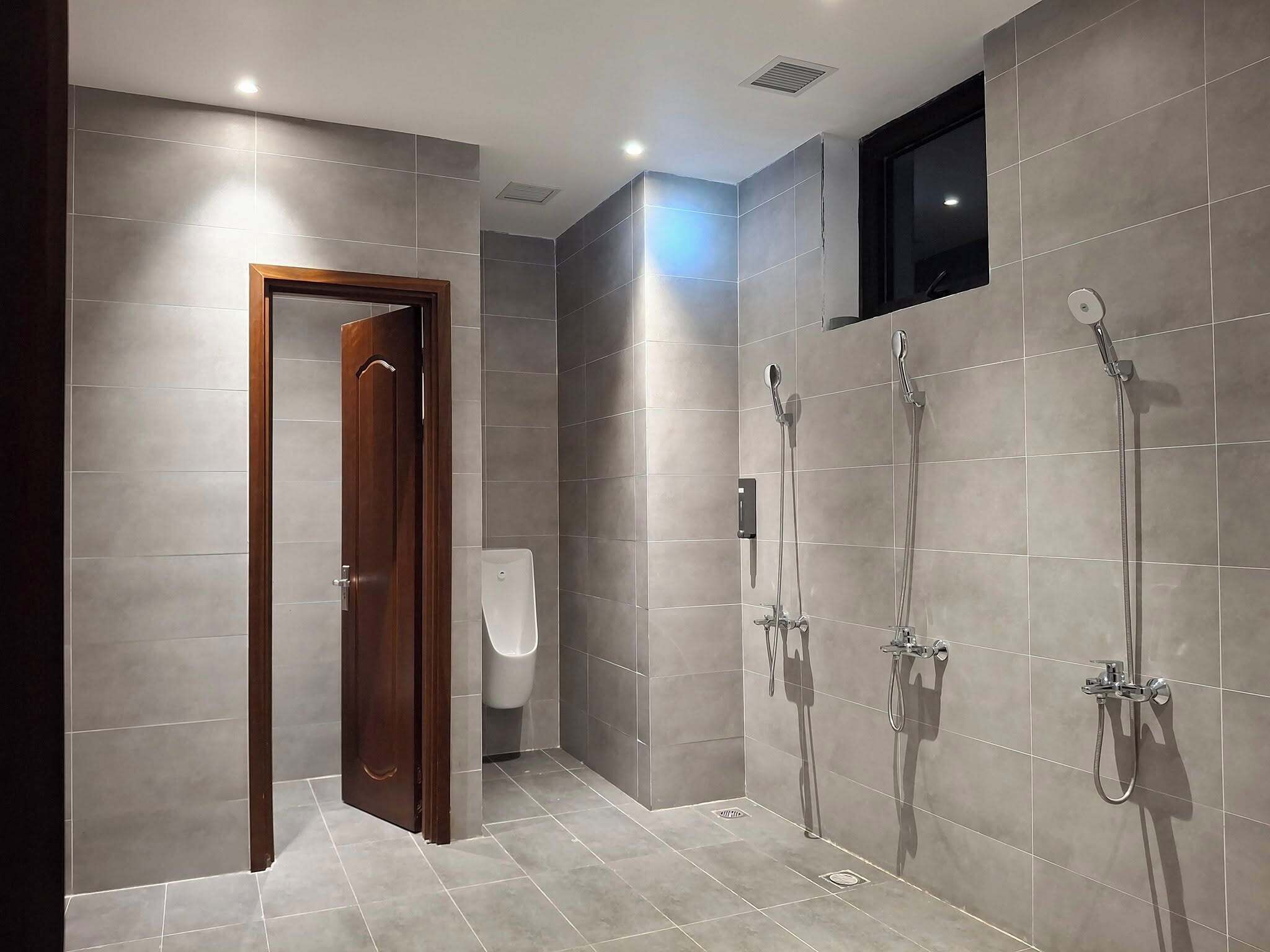 A modern bathroom with grey tiles, a wooden door, a urinal, and two showerheads.