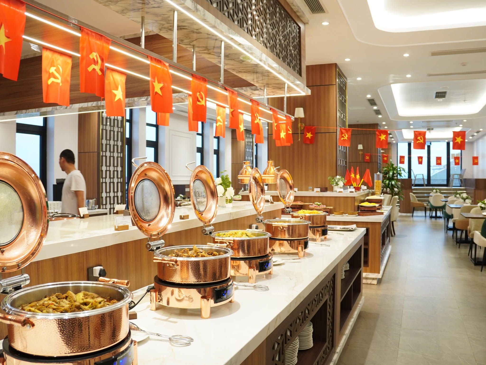 A buffet area with multiple food stations, a person in the background, and red flags hanging from the ceiling.