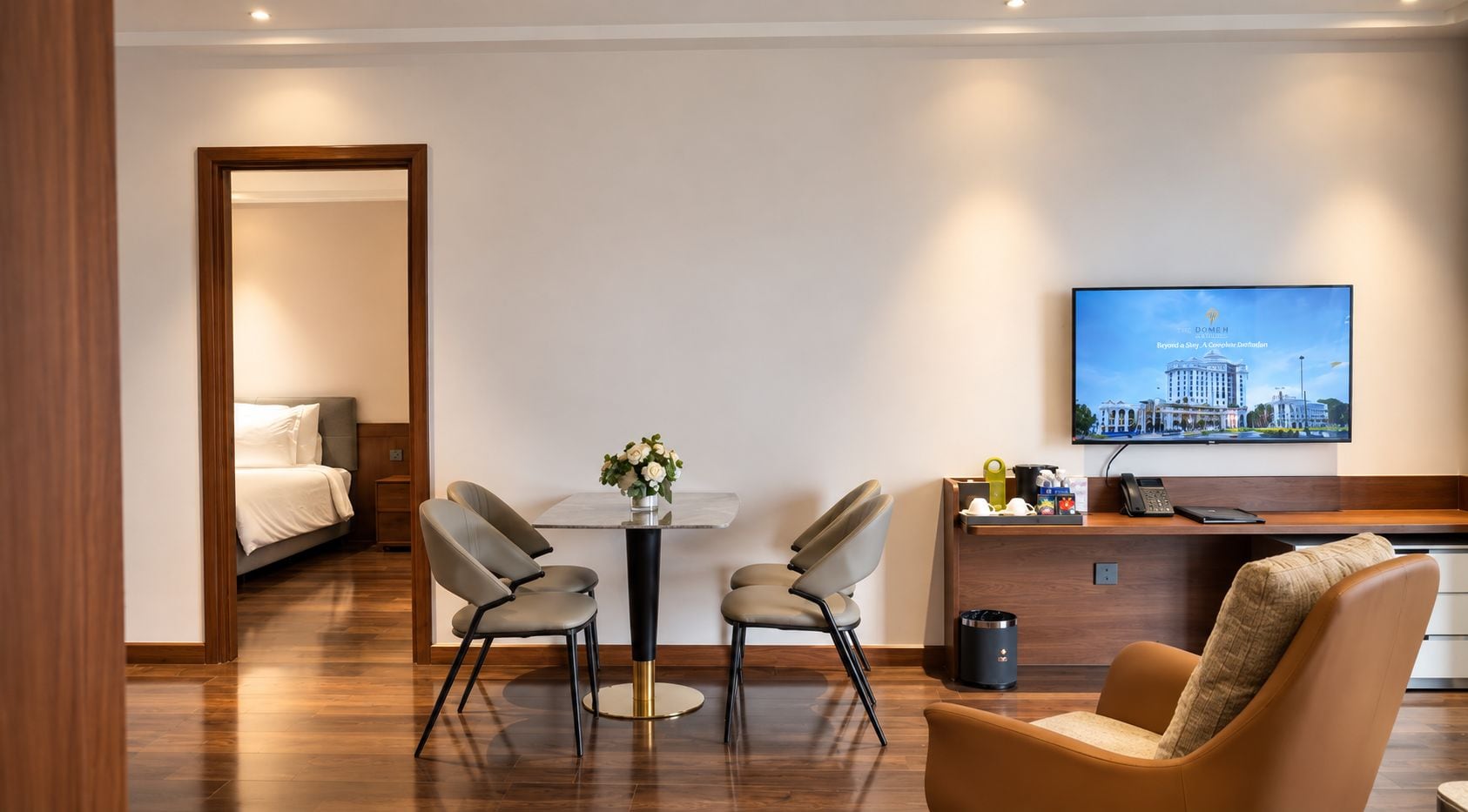 A hotel room with a bed, a desk with a computer, a TV, and a seating area with chairs.