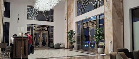 A spacious lobby with a high ceiling, marble flooring, and a grand chandelier. There are seating areas and a reception desk.
