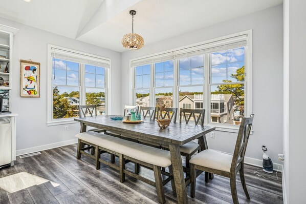 A dining area with a wooden table and chairs, large windows with a view of houses, and a ceiling light fixture. - 5 Bedroom Bayside Beach House! (South Bethany)
