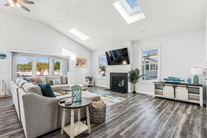 A spacious living room with a fireplace, a television mounted on the wall, and a large window offering a view of the outdoors. - 5 Bedroom Bayside Beach House! (South Bethany)