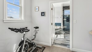 Interior de una vivienda con una bicicleta estática en una habitación con un ventanal grande, una puerta que da a una zona con baldosas y un cuadro pequeño en la pared.
