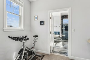 Interior de una vivienda con una bicicleta estática en una habitación con un ventanal grande, una puerta que da a una zona con baldosas y un cuadro pequeño en la pared.