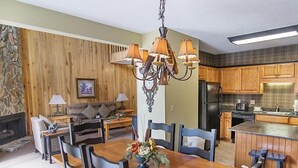 Un comedor con una mesa y sillas de madera, una araña y una cocina al fondo.