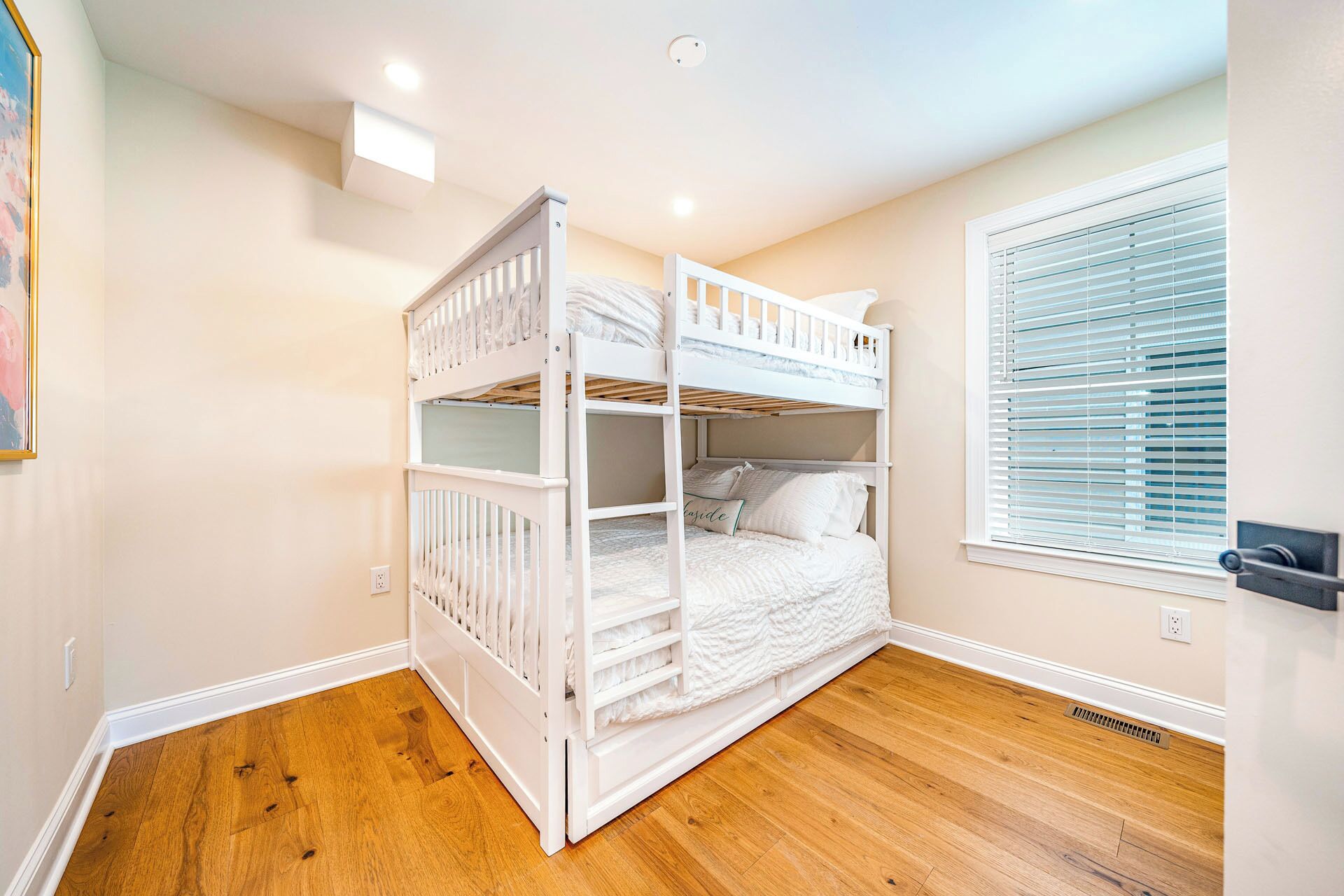 A room with a bunk bed, wooden flooring, a window with blinds, and a wall-mounted light fixture.