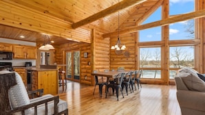 A log cabin interior with a dining area, kitchen, and large windows offering a view of the outdoors. - Fawn Lake Retreat | Pontoon, Beach & Game Room (Crosslake)