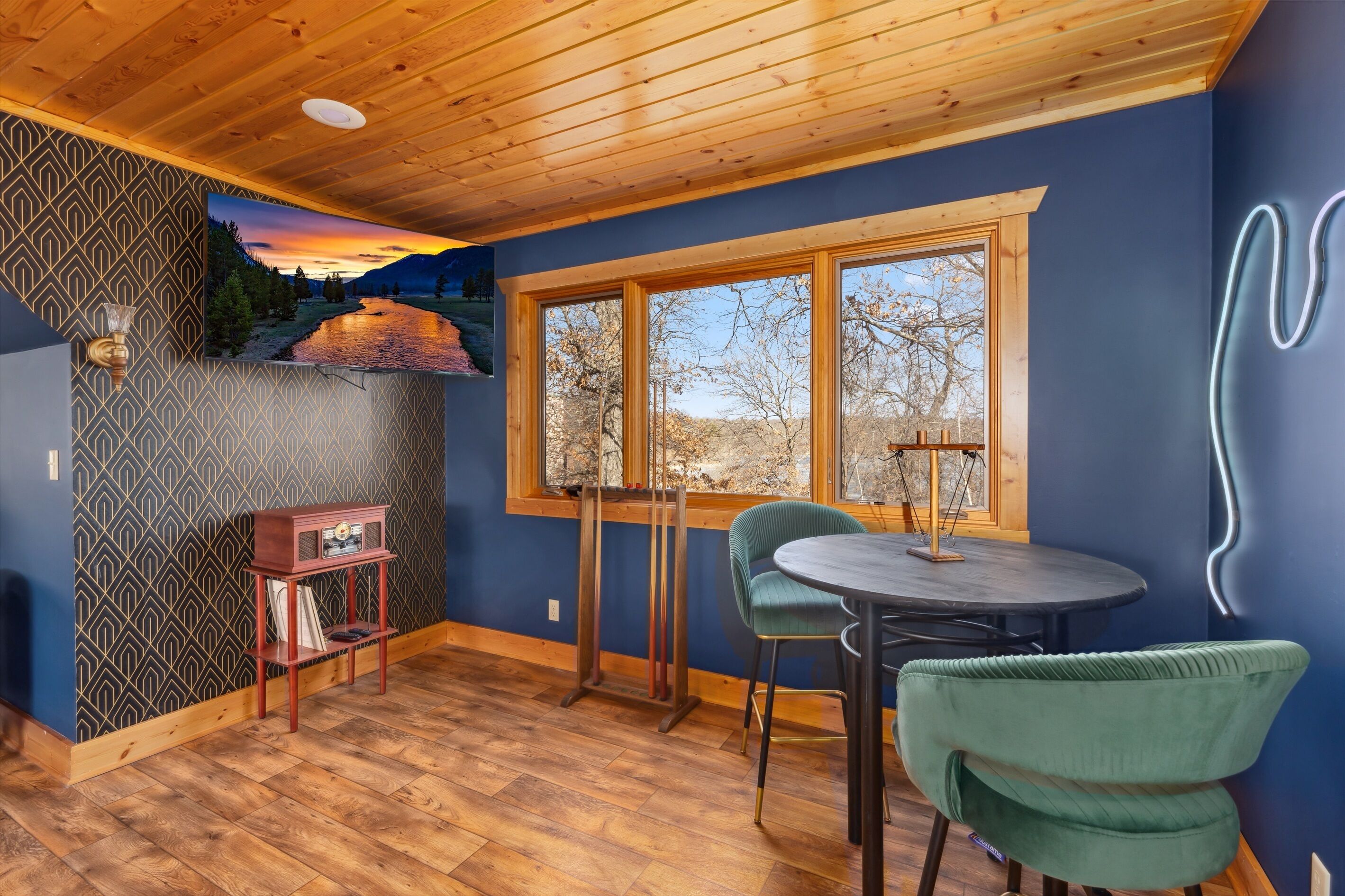 A room with a wooden ceiling, a large window with a view of trees and mountains, a table with chairs, and a radio on a shelf.