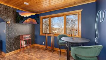 A room with a wooden ceiling, a large window with a view of trees and mountains, a table with chairs, and a radio on a shelf.