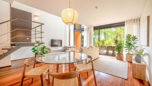 A modern living room with a glass dining table, wicker chairs, a staircase with a geometric design, and a view of a garden through large windows.