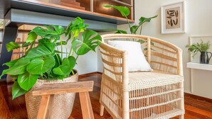A wicker chair with a white cushion, a woven side table, and large green plants in a woven basket.