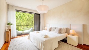 A modern bedroom with a large bed, a balcony with a view of greenery, and a white color scheme.