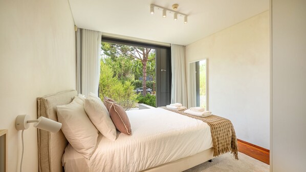 A bedroom with a large bed, a window with curtains, and a view of greenery outside.