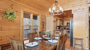 Interior de una cabaña de madera con una mesa de comedor dispuesta para cuatro personas, una araña y una zona de cocina con refrigerador y estufa.