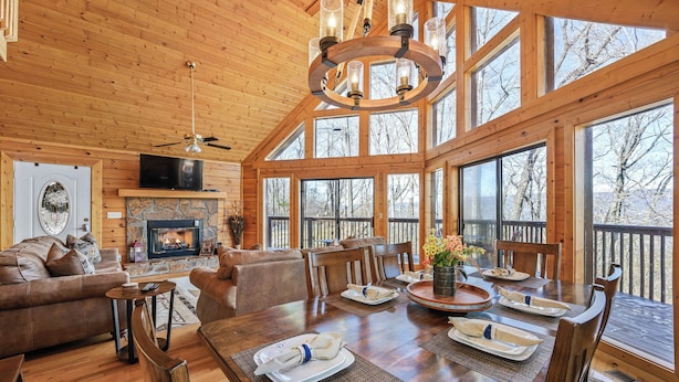 Una cabaña de madera espaciosa con comedor, chimenea y ventanas amplias que ofrecen vistas de los árboles circundantes.