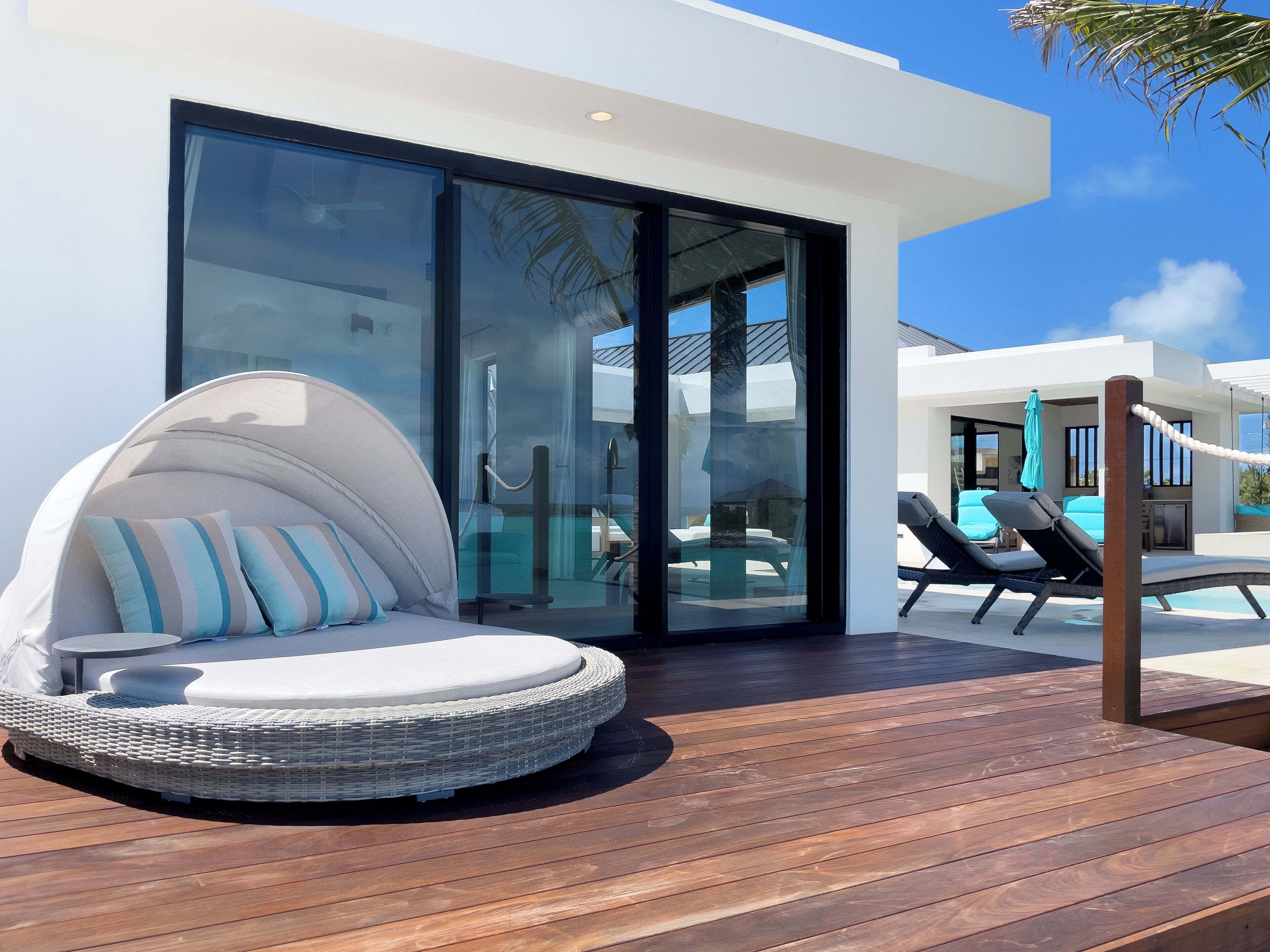 Un espace extérieur moderne aménagé avec des sièges, notamment un fauteuil en osier rond doté de coussins, une terrasse en bois et une porte en verre donnant accès à une zone piscine.