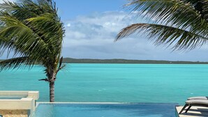Una piscina infinita con dos sillas blancas y una mesa, con vista al mar turquesa y palmeras.