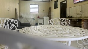Un comedor con una mesa redonda y sillas blancas, un televisor montado en la pared, y una cocina al fondo.