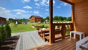 Una terraza de madera con mesas de picnic y bancos, vista a un parque infantil y una casa al fondo.