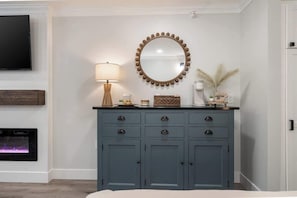 A room with a grey dresser, a round mirror with a bead trim, a wall-mounted TV, and a fireplace.