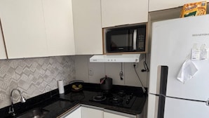 A compact kitchen with white cabinets, a black countertop, a microwave, a refrigerator, and a water dispenser. - Beach House in Barra, Ilhéus (Barra do Itaípe Ilhéus)