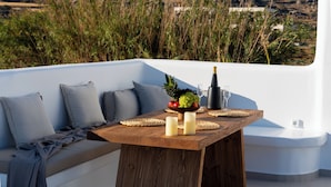 Una mesa de madera con un frutero, velas y una botella de champán en un balcón con un sofá blanco y cojines grises.
