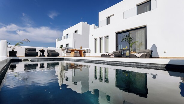 Un área de piscina moderna al aire libre con un cielo azul despejado, un edificio blanco y mobiliario exterior.