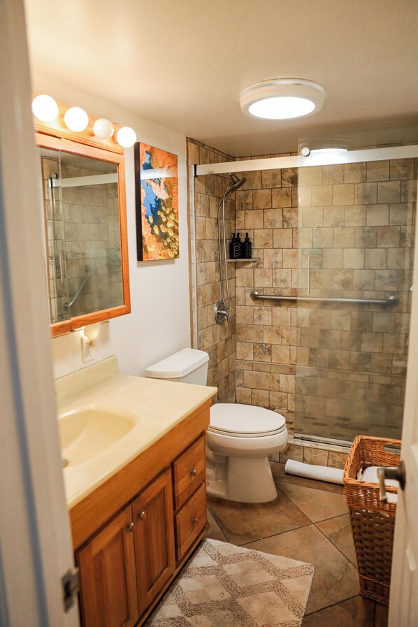 Un baño con tocador de madera, inodoro blanco, ducha con cerramiento de vidrio y suelo de baldosas.
