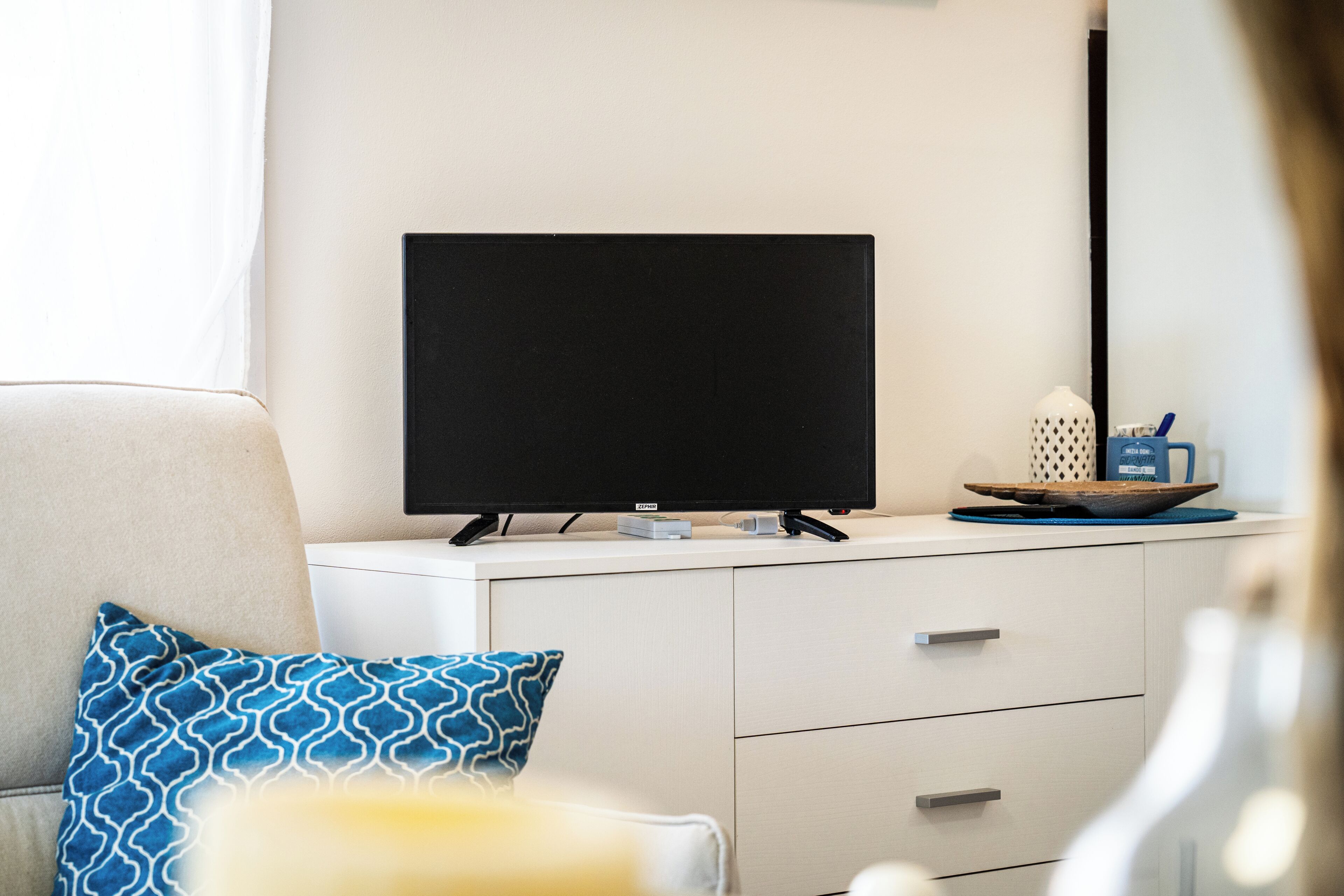 Un salon moderne avec un téléviseur à écran plat posé sur un meuble blanc, un canapé agrémenté de coussins à motifs et une table d’appoint sur laquelle est placé un objet décoratif.