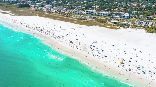 An aerial view of a beach with a long stretch of white sand, clear turquoise water, and a dense urban area in the background.