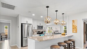 A modern kitchen with stainless steel appliances, a large island with bar stools, pendant lighting, and a view of the outdoors.