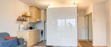 A modern apartment with a grey sofa, a white ottoman, a kitchen with white cabinets, and a large white wardrobe.