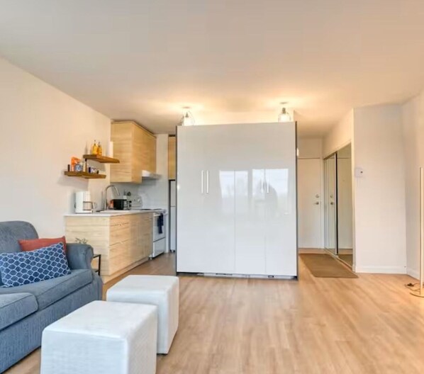 Un apartamento moderno con un sofá gris, un reposapiés blanco, una cocina con armarios blancos y un amplio armario blanco.