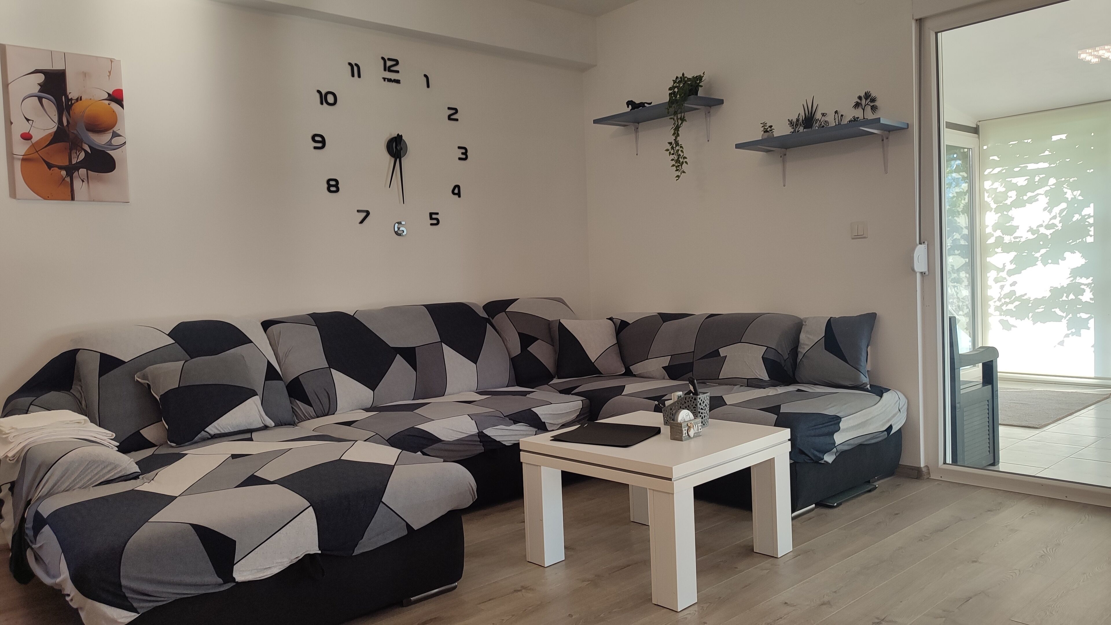 Un salon moderne avec un canapé d’angle, une table basse blanche et une horloge murale. On y trouve deux étagères murales avec des plantes et une porte donnant sur un espace extérieur.