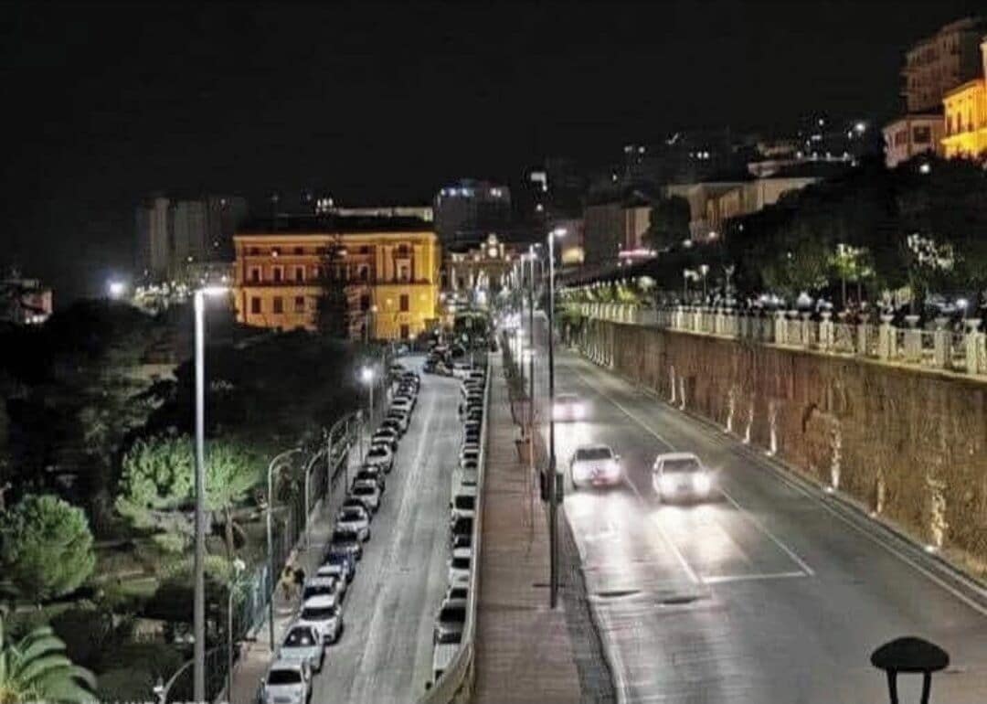 Rua urbana noturna com carros estacionados, postes de luz e edifícios.