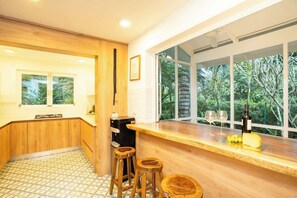 A kitchen with wooden cabinets, a countertop, stools, and a window with a view of greenery.