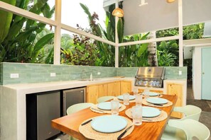 A kitchen with a dining area, a grill, and a view of lush greenery. - Ocean Gardens: Private Retreat, Private Pool & BBQ (Esterillos Oeste)