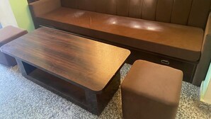Un sofá de cuero marrón con un banco a juego y una mesa de centro de madera en una habitación con pared verde y una ventana con cortina translúcida.