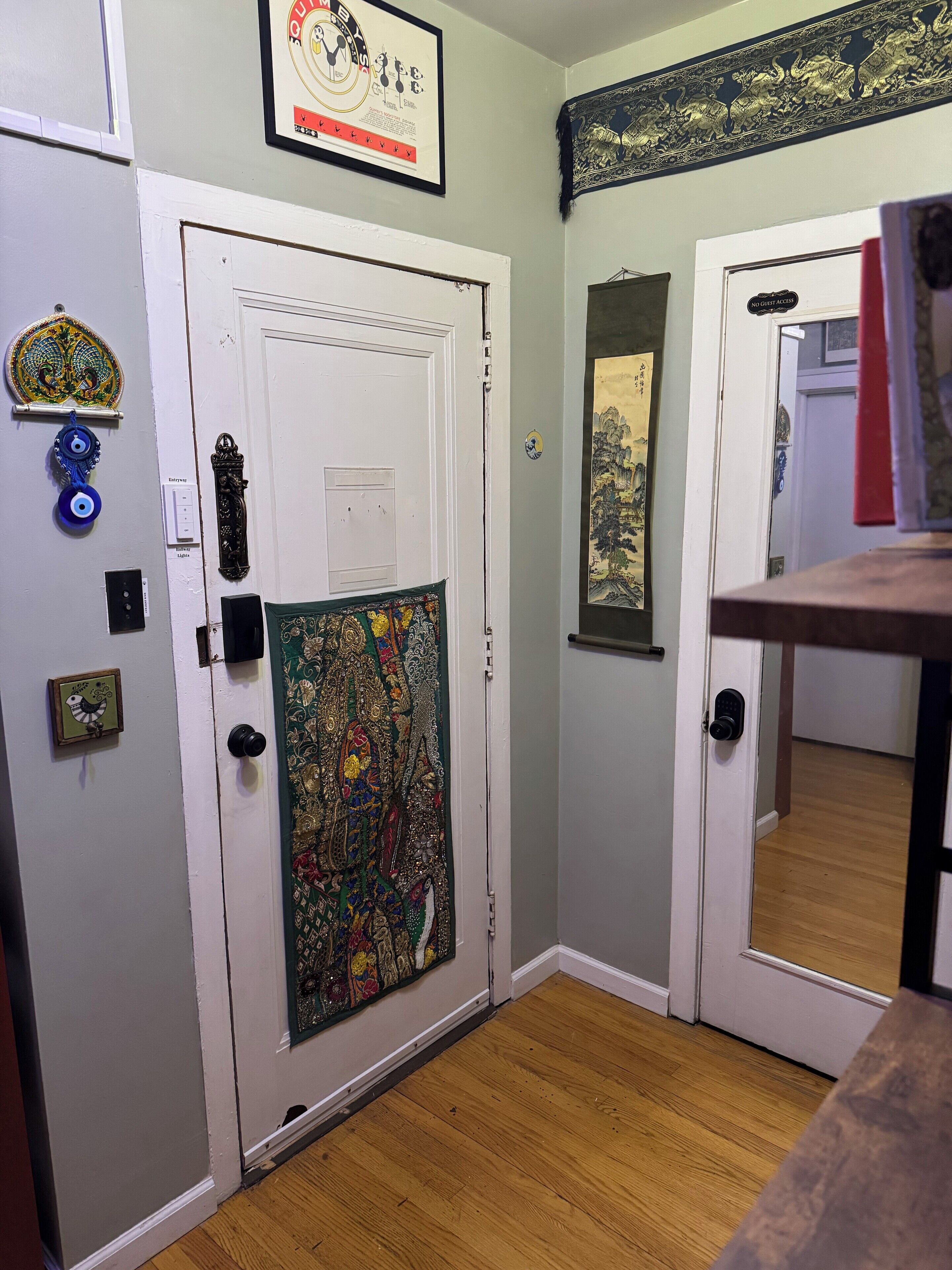 A hallway with two white doors, one with a colorful tapestry and the other with a framed picture. There are decorative plates on the wall and a small framed picture on the door.