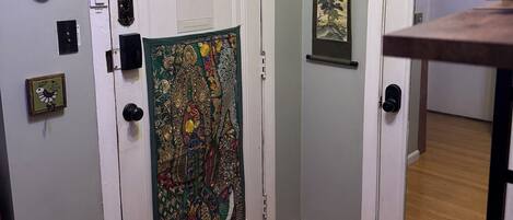 A hallway with two white doors, one with a colorful tapestry and the other with a framed picture. There are decorative plates on the wall and a small framed picture on the door.