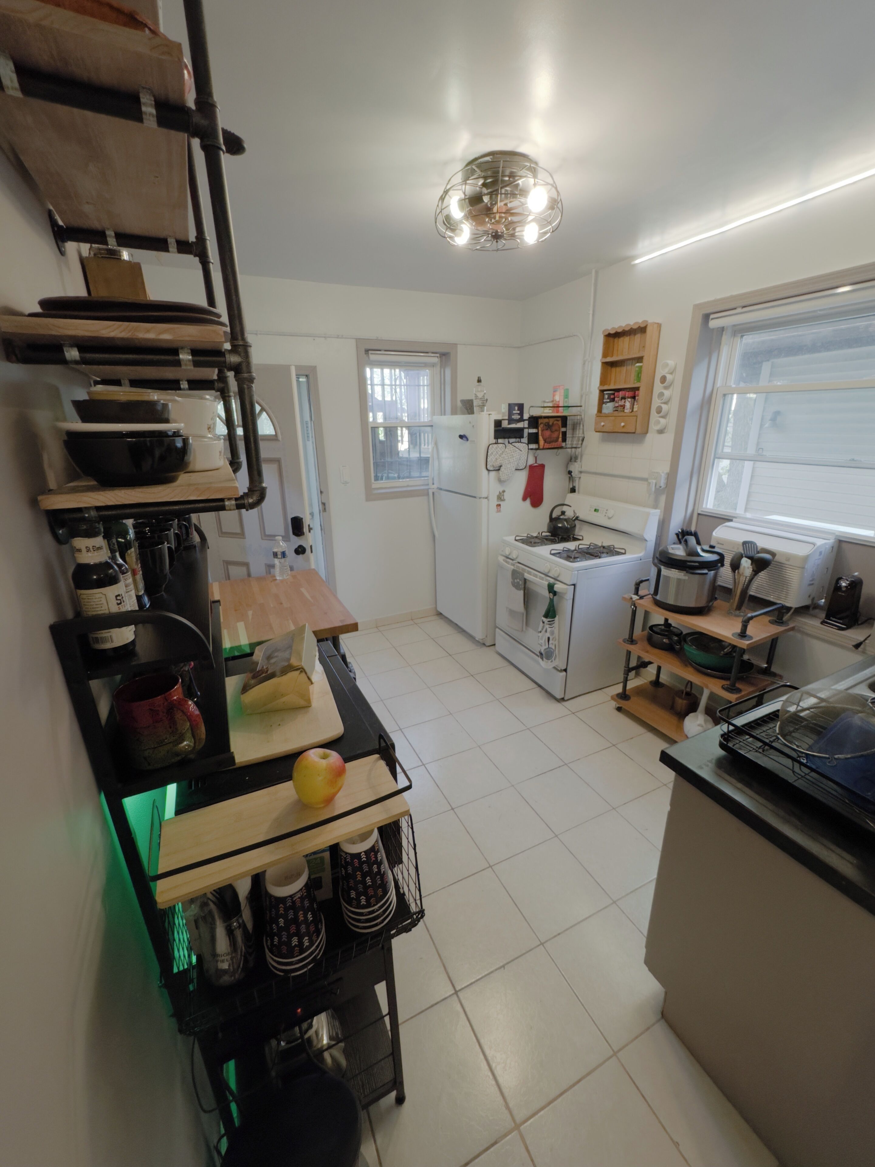 Une cuisine compacte avec des meubles blancs, une cuisinière encastrée et un réfrigérateur. On y trouve une étagère en bois avec de la vaisselle et une planche à découper sur laquelle se trouve une pomme. Une petite fenêtre laisse entrer la lumière naturelle.