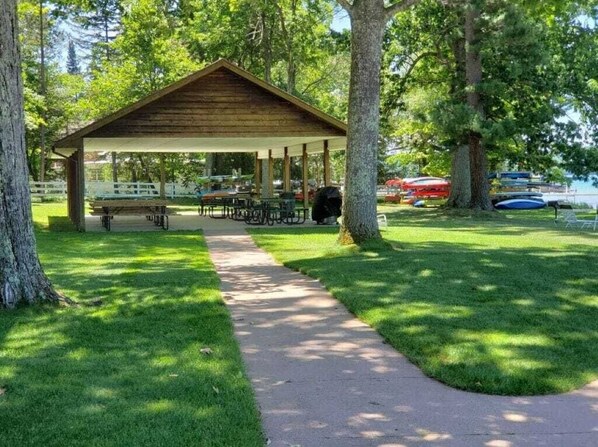 Un área de picnic techada con mesas y bancos, rodeada de árboles y un sendero.