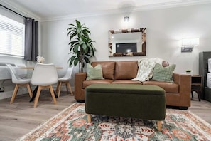 Una sala de estar con un sofá marrón, un otoman verde, un tapete estampado, un espejo grande y una planta en maceta.