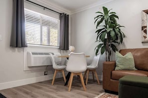 Una sala de estar con un sofá de cuero marrón, una mesa de comedor con dos sillas, una planta verde grande y una ventana con persianas.