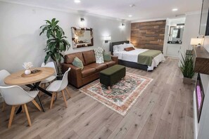 Una habitación de hotel moderna con piso de madera, un sofá de cuero marrón, una mesa de comedor redonda, un reposapiés verde, una cama con ropa blanca y un pequeño macetero.
