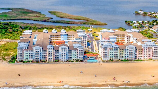 A beachfront hotel with multiple-story buildings, a sandy beach, and a view of the ocean and marshy areas. - New! Island Breeze/3Bd/Oceanfront/ Beach/Pool/Gym (Virginia Beach)