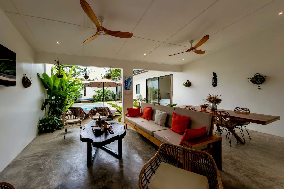 A modern living space with a sofa, dining table, and chairs, a ceiling fan, and a view of a pool and greenery outside.